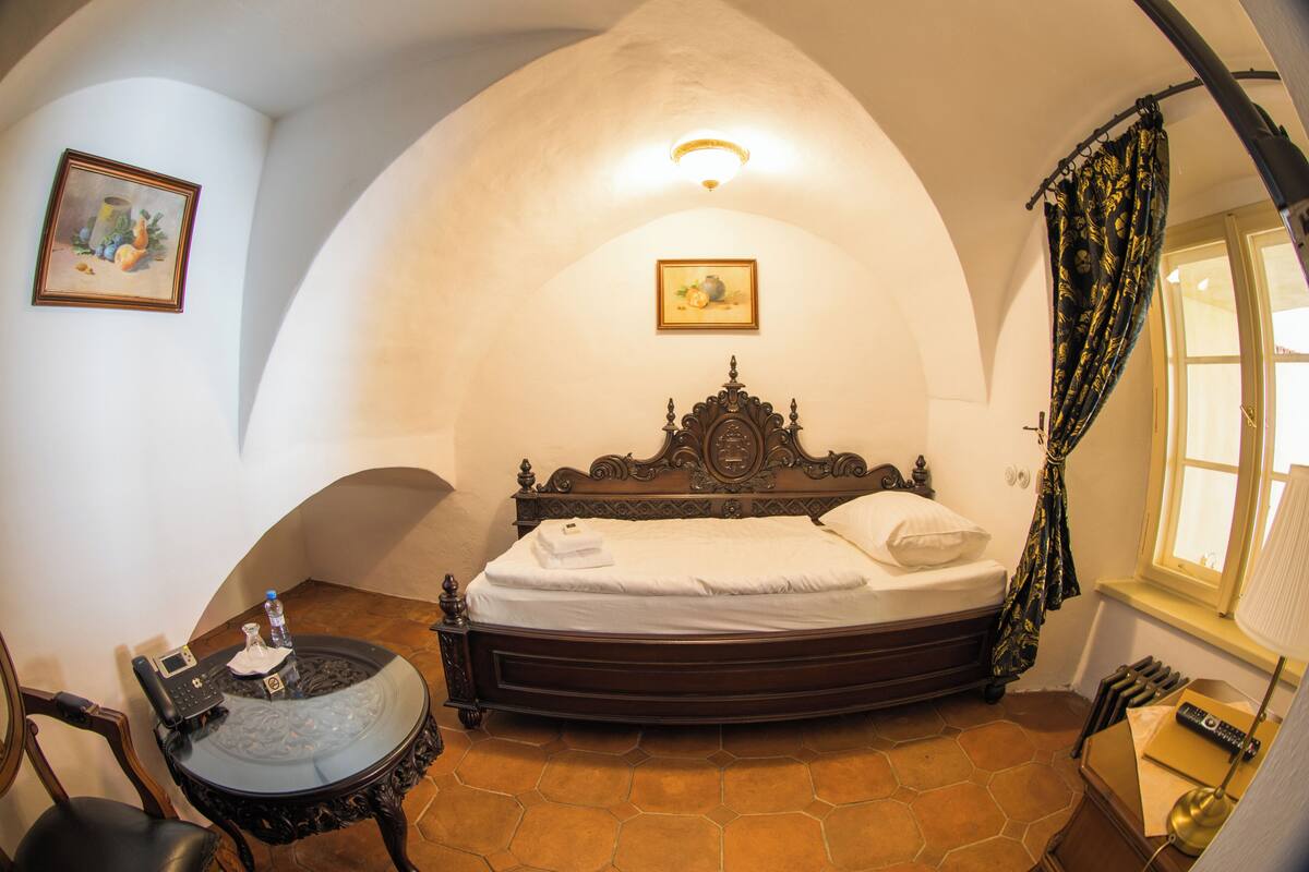 A room with a bed, a chair, a table, a lamp, a window with curtains, and two paintings on the wall.