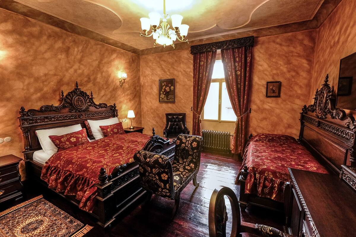 A traditional room with a large bed, a smaller bed, a chair, a table, and a chandelier.