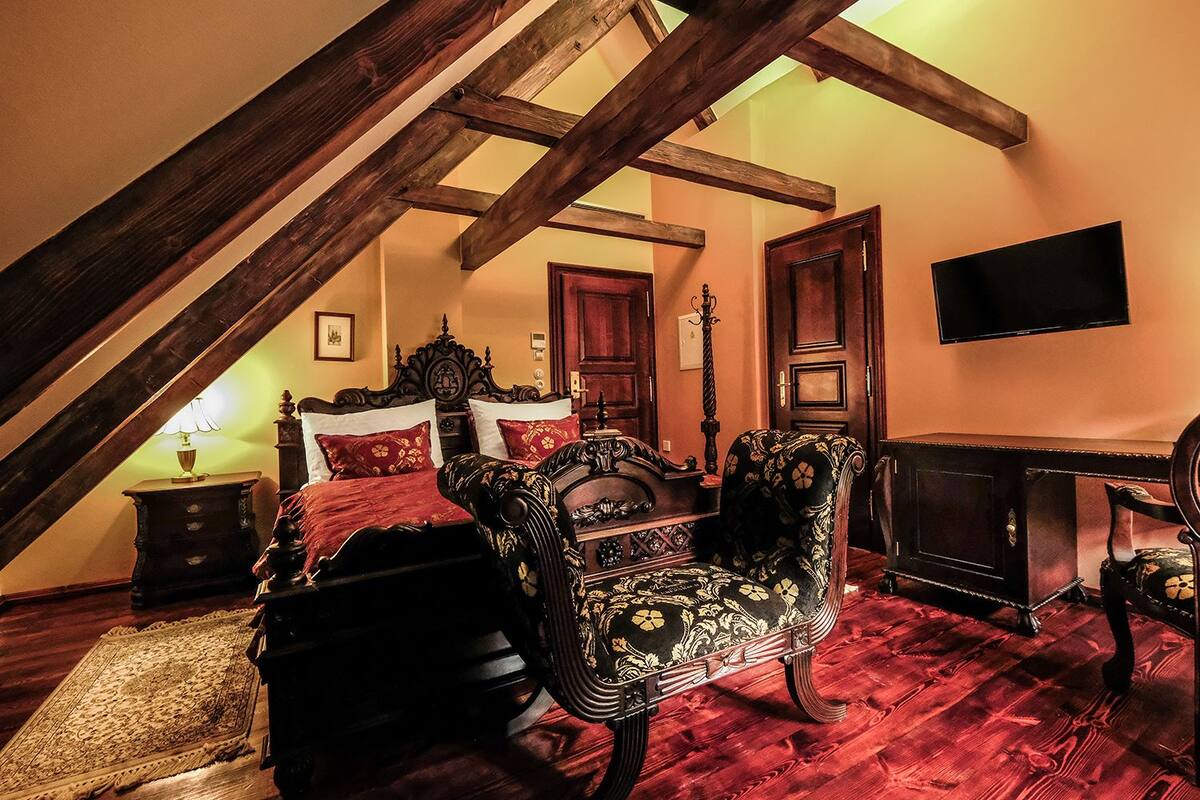 A bedroom with a sloped ceiling, a bed with red bedding, two ornate chairs, a wooden desk, and a television on the wall.