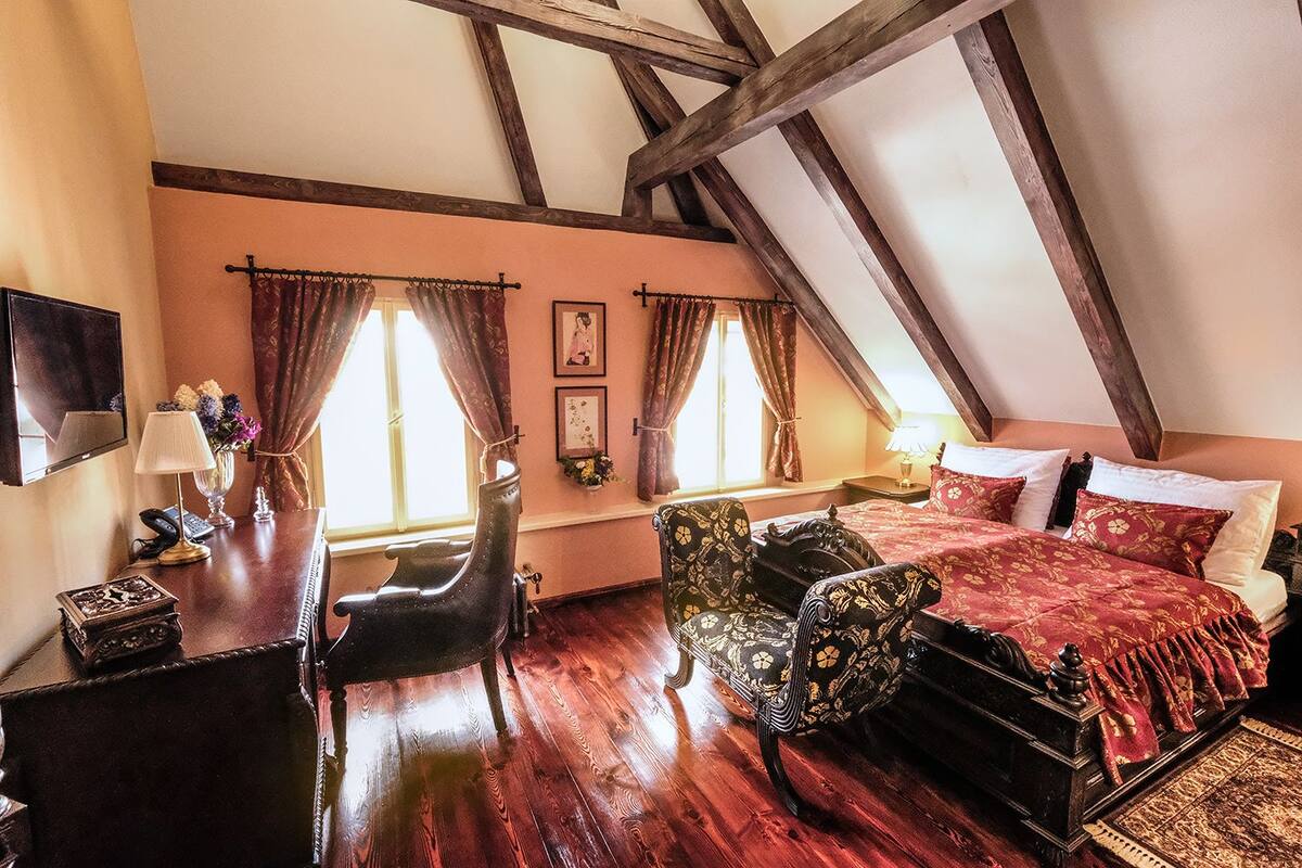 A bedroom with a wooden ceiling, a bed with red bedding, a desk with a lamp, and a chair.