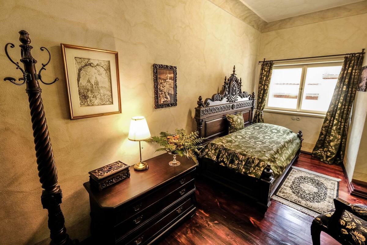 A bedroom with a bed, a nightstand with a lamp, a dresser, and a window with curtains.