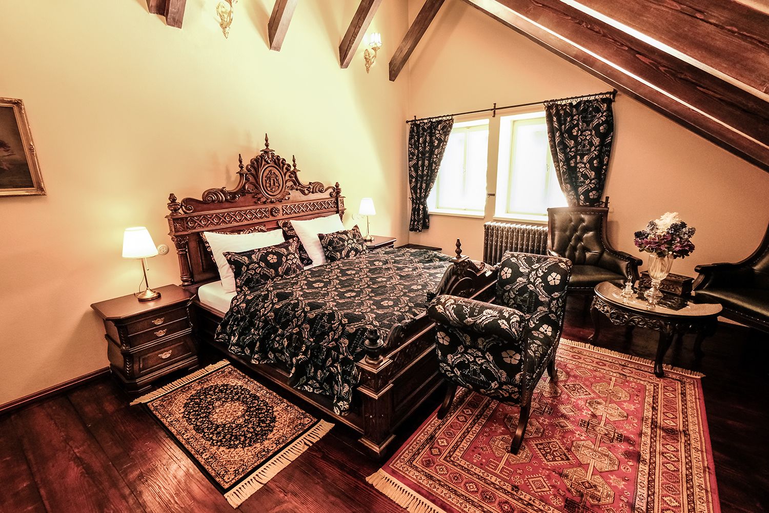 Un dormitorio con una cama grande, una mesita de noche, una silla, una mesa con un florero y una ventana con cortinas.
