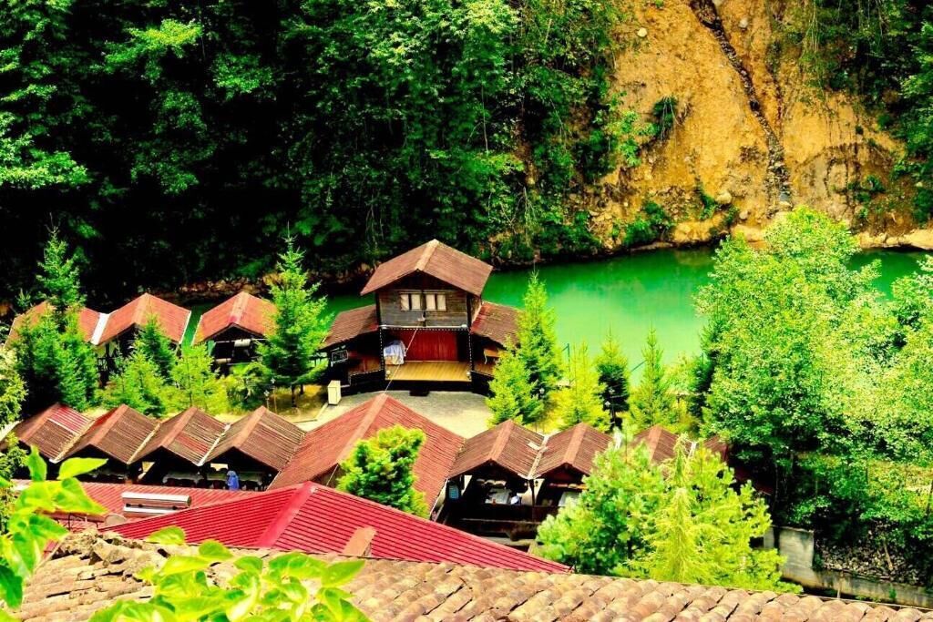 A cluster of traditional wooden cabins with sloped roofs, surrounded by dense forest and a clear green lake.