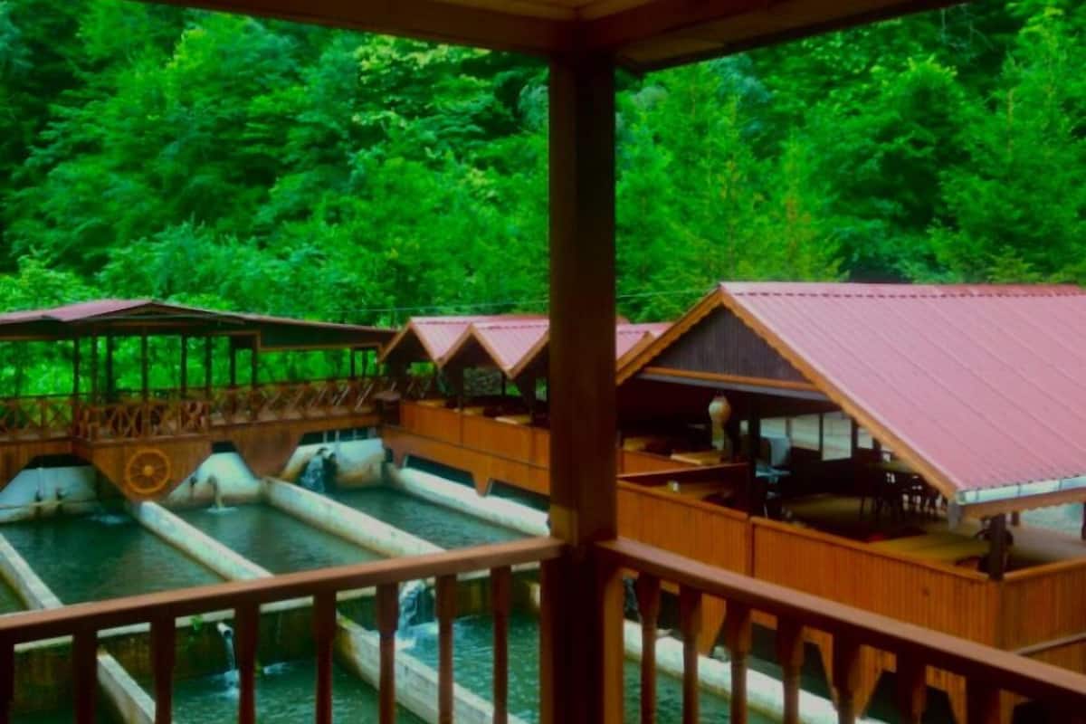 A wooden deck with a view of a river and a bridge, wooden cabins with red roofs, and a wooden railing.