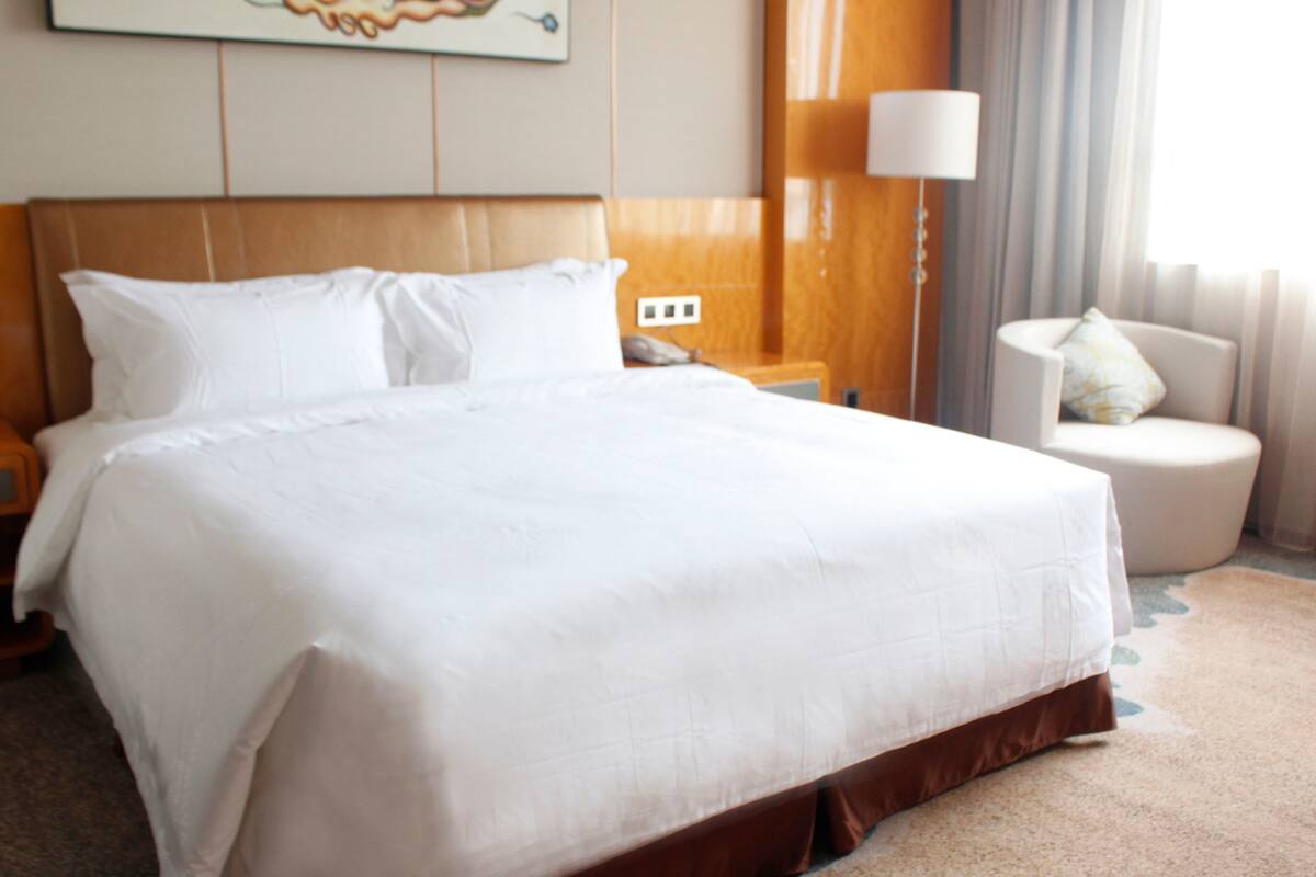 A large bed with white linens, a wooden headboard, a bedside table with a lamp, and a chair with a cushion.