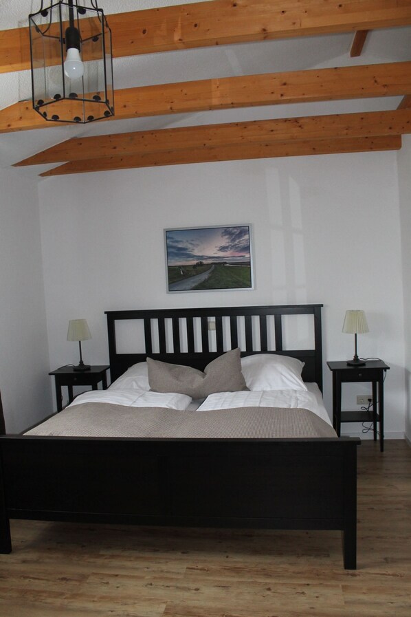 A bedroom with a wooden bed frame, two bedside tables with lamps, a framed picture on the wall, and a wooden ceiling beam. - Zum alten Krug (Wangerland)