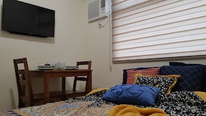 A bedroom with a bed, a table with a cup and saucer, a chair, a TV mounted on the wall, and a window with blinds. - Aorta Your Home in The Heart of EDSA (Mandaluyong)