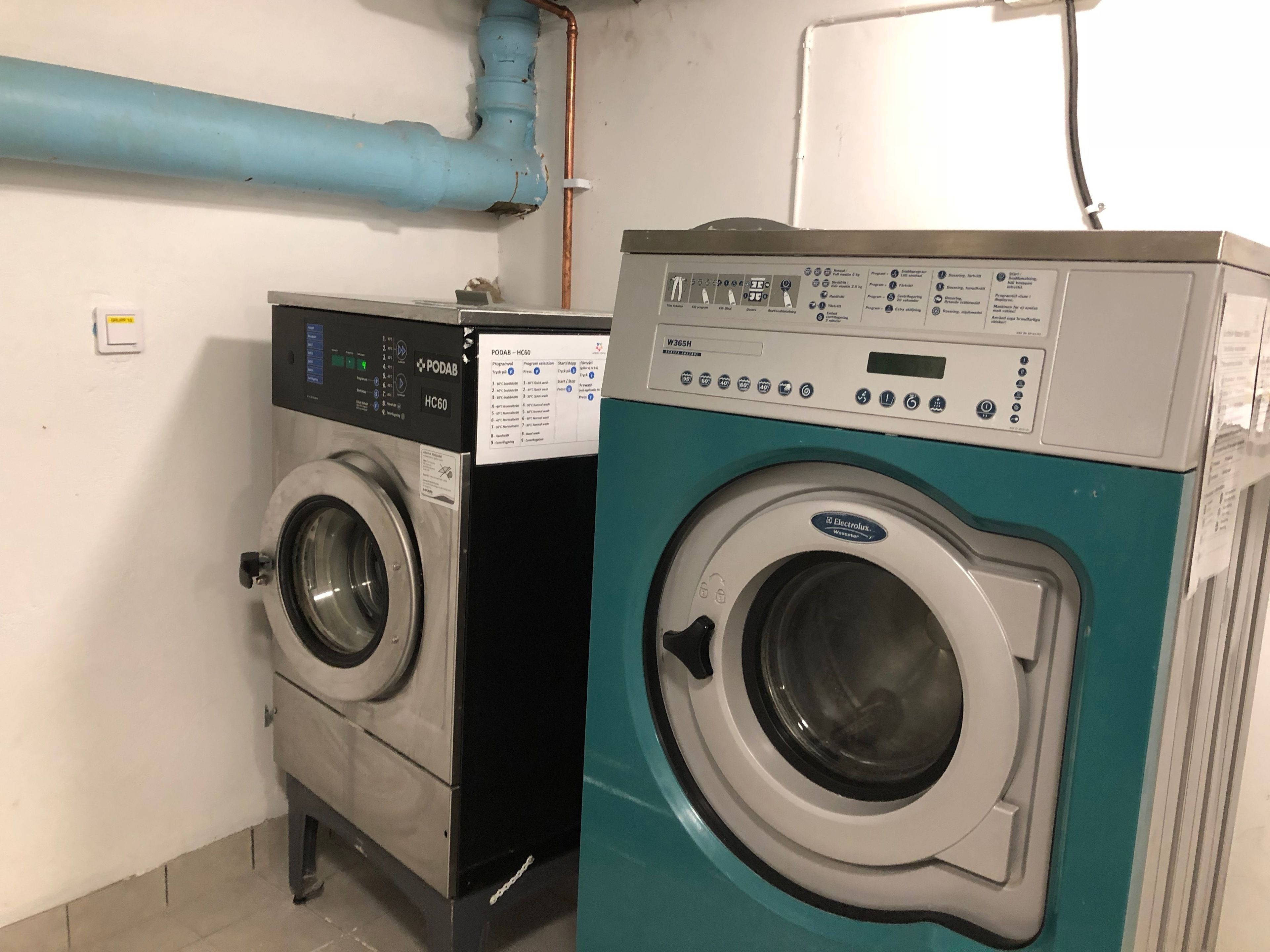 Two front-loading washing machines in a laundry room with a visible control panel and a blue pipe on the wall.