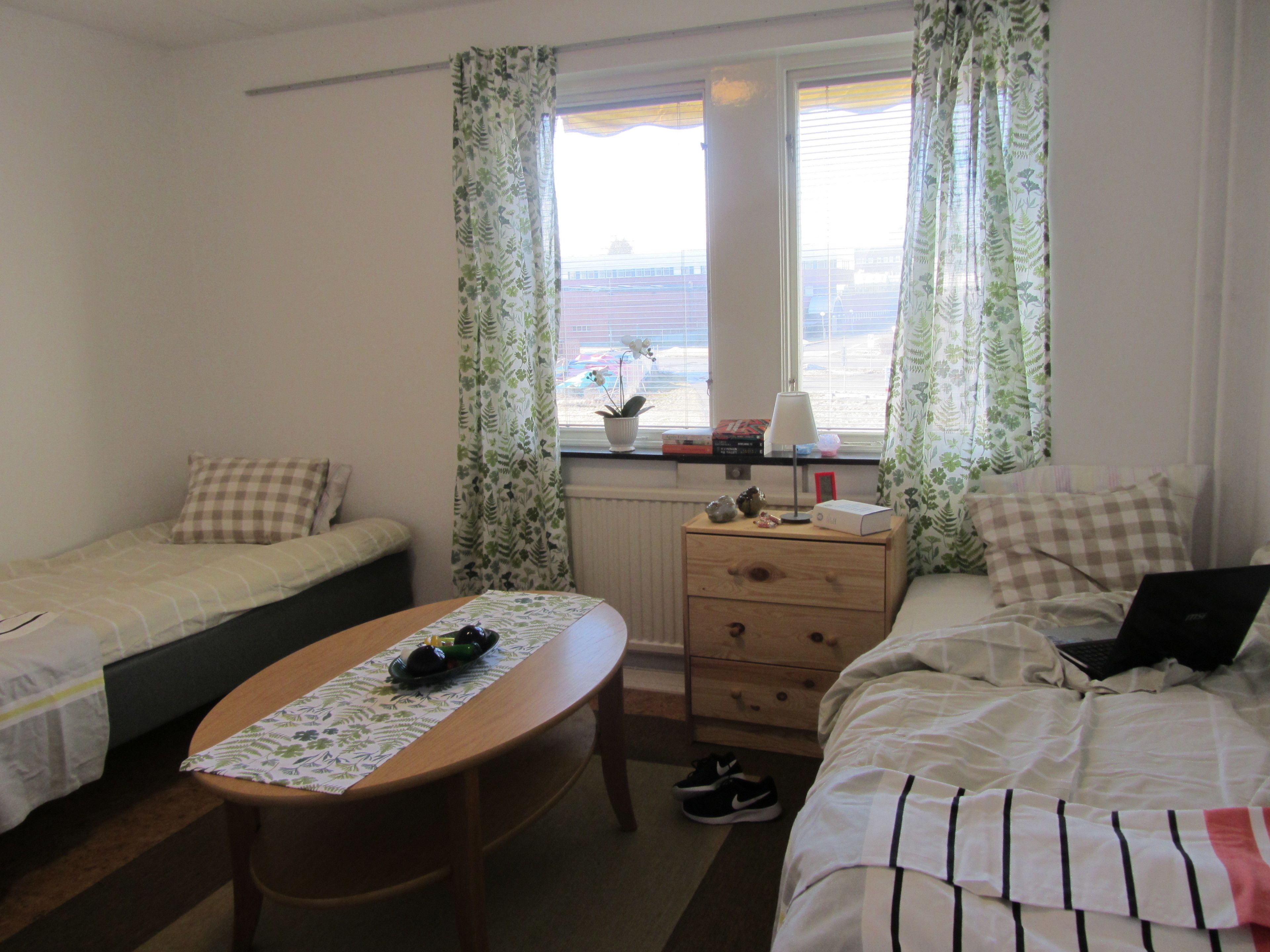 A bedroom with two single beds, a wooden table, a dresser, and a window with curtains.