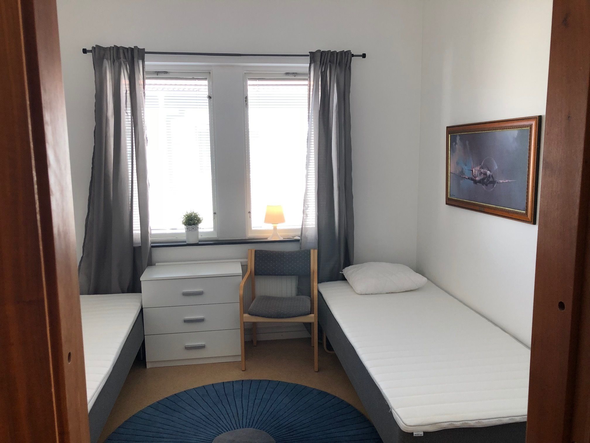 Une petite pièce, bien rangée, avec deux lits simples, un bureau avec des tiroirs, une chaise, une petite table avec une lampe et un tableau encadré au mur.