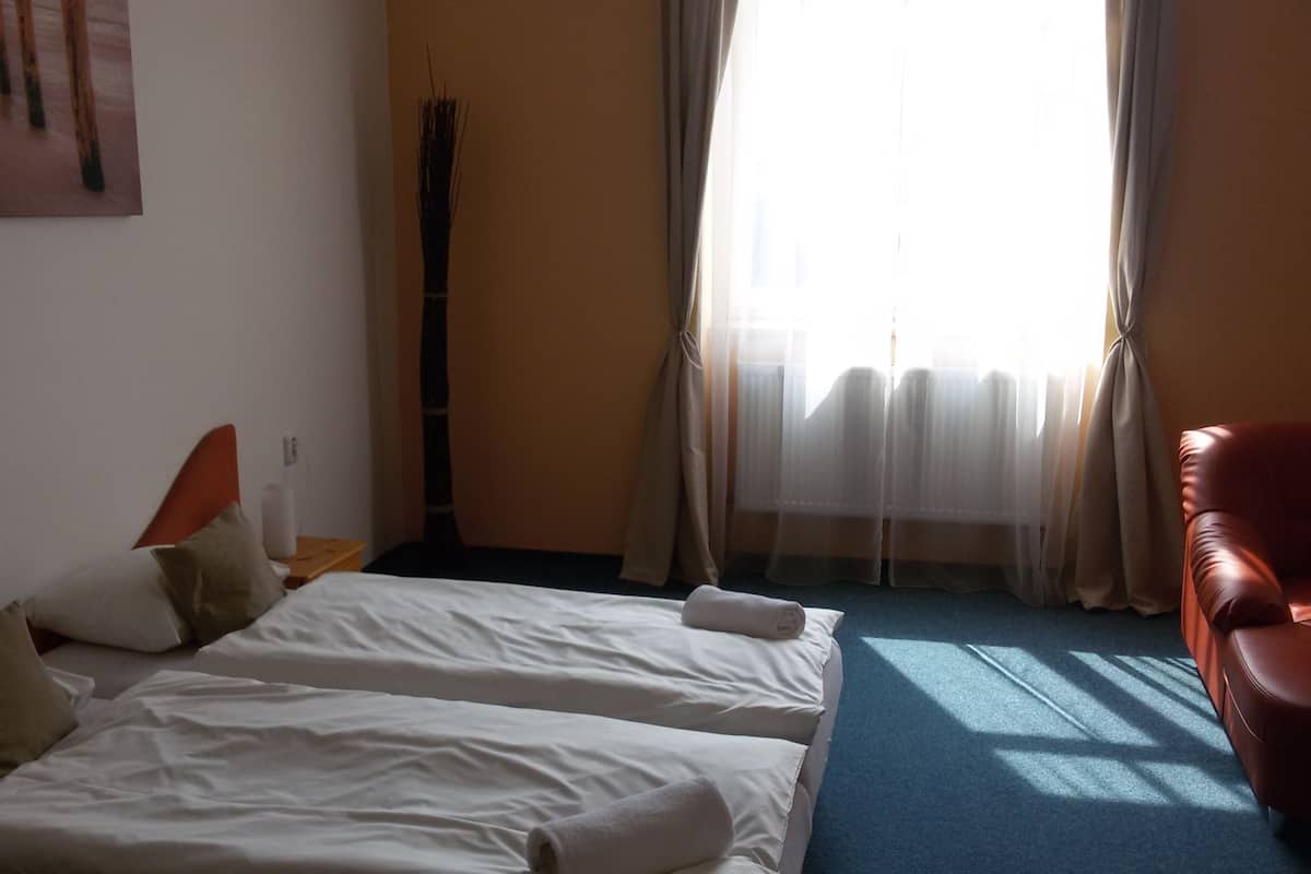 Habitación de hotel con dos camas, un sillón de cuero rojo, un ventilador de pie y una ventana con cortinas traslúcidas.