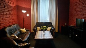 Una sala de estar con pared de ladrillo, un sofá de cuero, una mesa de centro con un jarrón de flores, un televisor y una lámpara de pie.