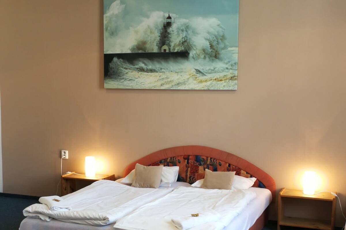 Una habitación de hotel con una cama grande, dos mesitas de noche con lámparas, un sillón tapizado en rojo y un cuadro de un faro en la pared.
