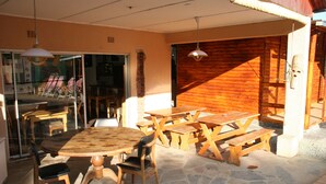 Um pátio externo com mesas e bancos de madeira, piso de pedra e uma porta de vidro que dá para um espaço interno.