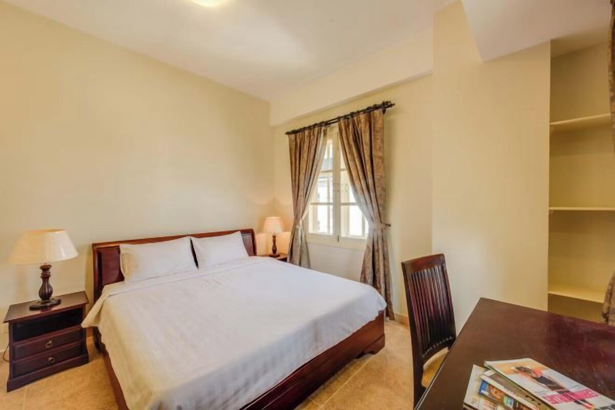 A bedroom with a bed, bedside tables, a window with curtains, a wooden chair, and a table with a magazine.