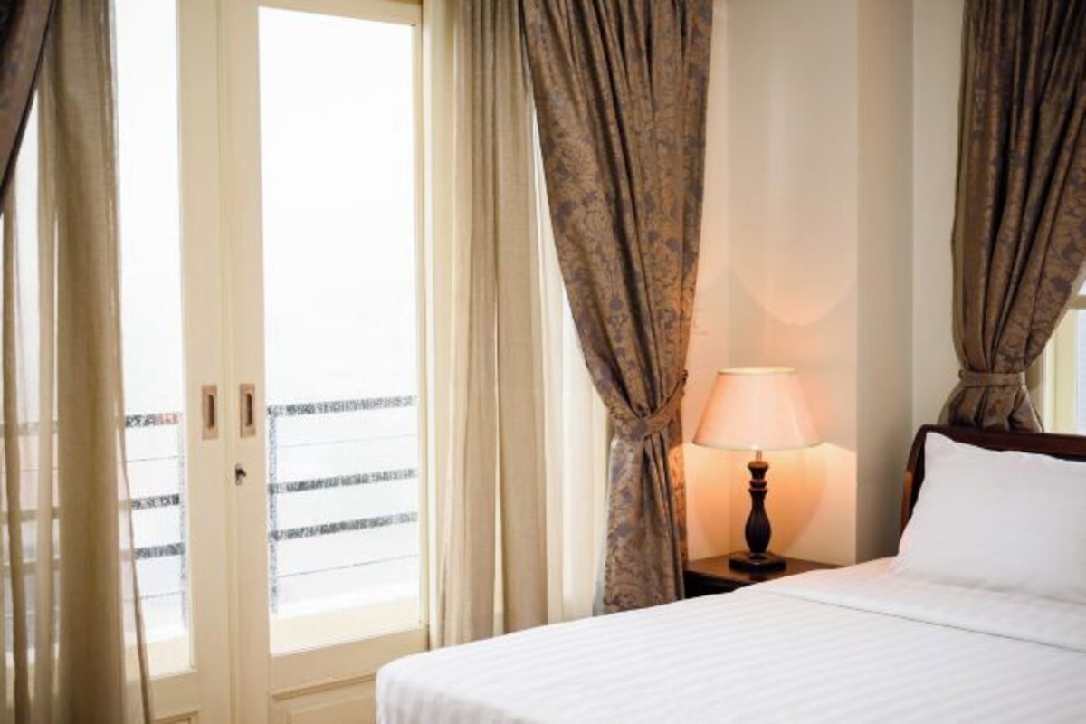 A hotel room with a bed, a nightstand with a lamp, and a view through a window with curtains.