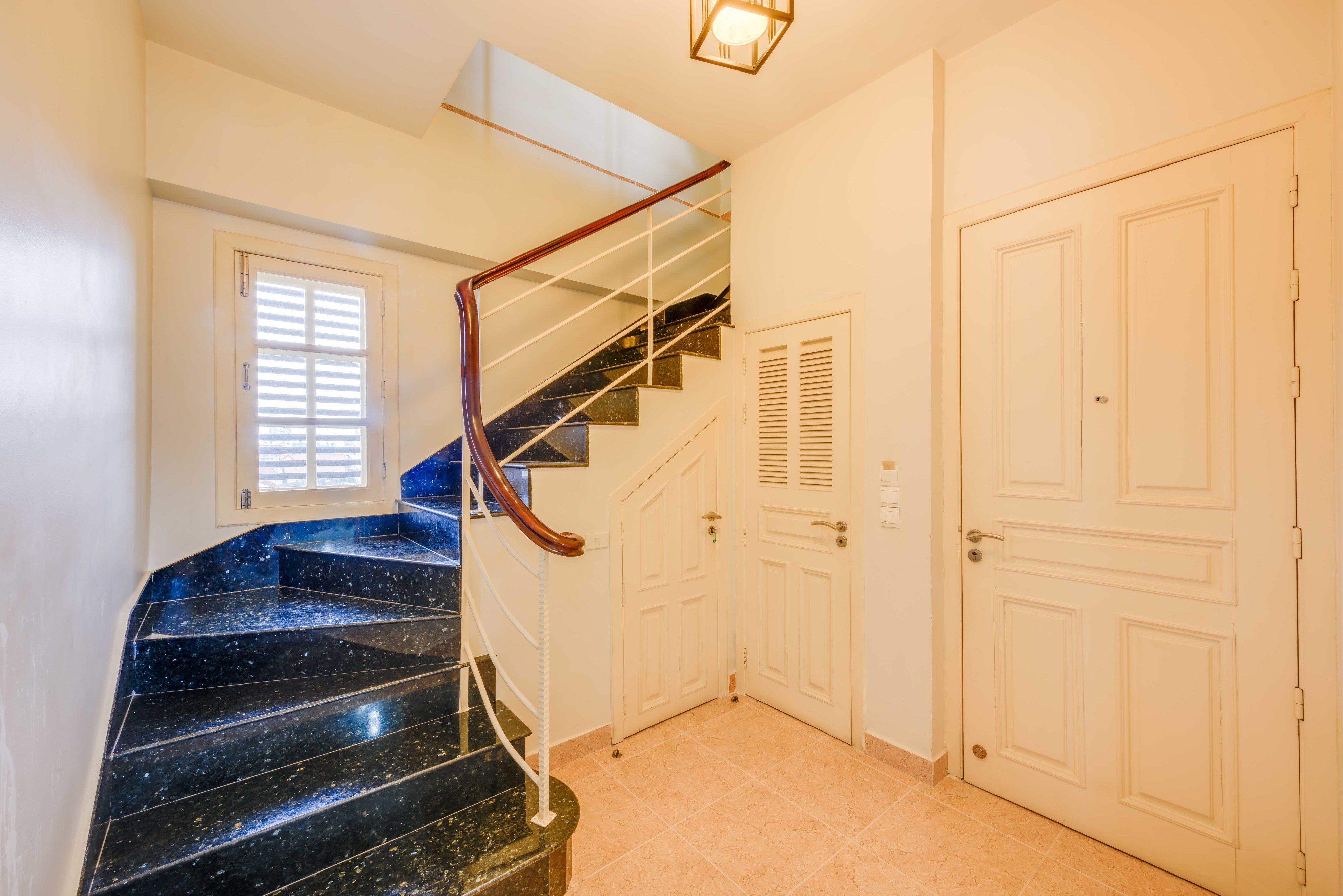 A staircase with black marble steps and a wooden handrail, leading to a landing with a white door and a window with blinds.
