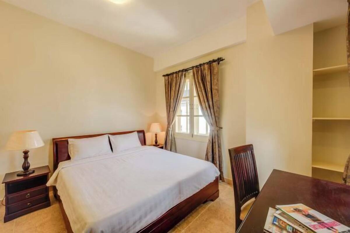 A bedroom with a bed, bedside tables, a chair, a window with curtains, and a wooden table with magazines.