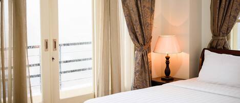 Habitación de hotel con cama, mesita de noche con lámpara y vistas a través de una ventana con cortinas.