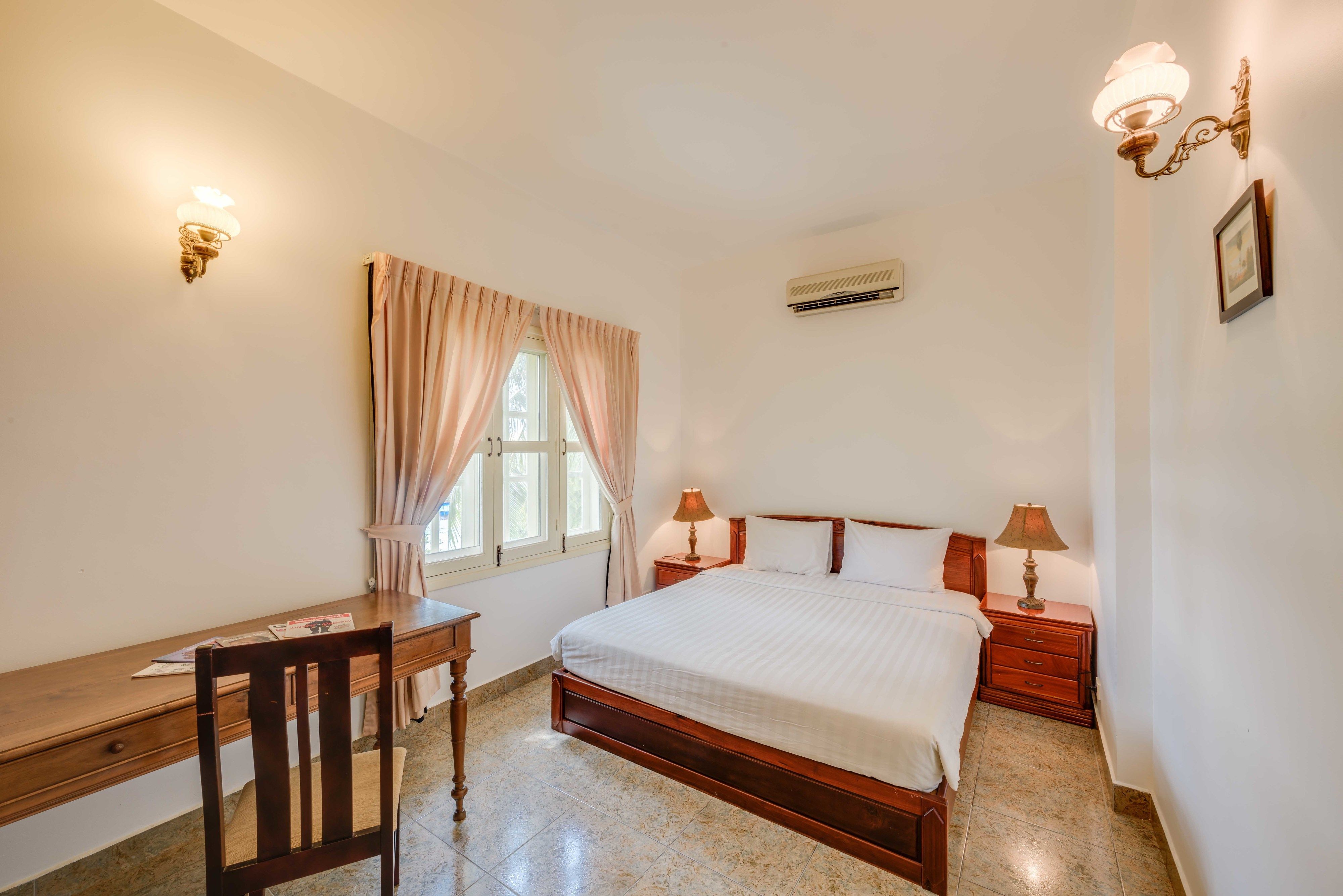 Quarto com cama de madeira, uma escrivaninha, uma cadeira, duas luminárias de cabeceira, uma janela com cortinas e um ar condicionado.