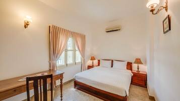 Quarto com cama de madeira, uma escrivaninha, uma cadeira, duas luminárias de cabeceira, uma janela com cortinas e um ar condicionado.