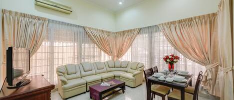 Una sala de estar con un sofá seccional color beige, una mesa de comedor lista para cuatro personas y un televisor sobre un mueble de madera.