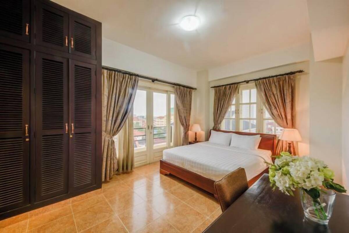 A hotel room with a bed, curtains, a nightstand, a vase with flowers, and a view through the window.