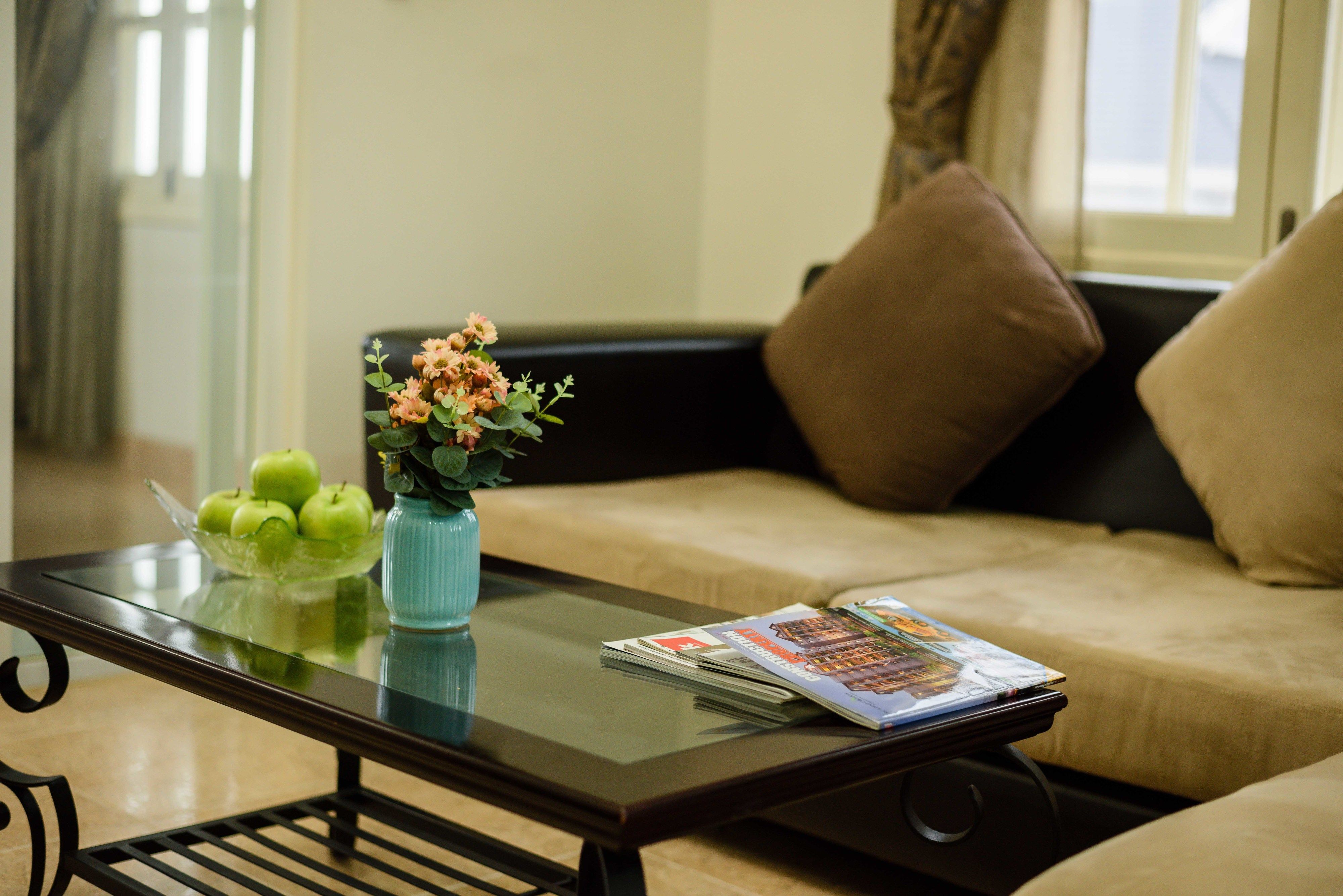 A living room with a glass coffee table, a bowl of green apples, a vase with flowers, and a magazine.