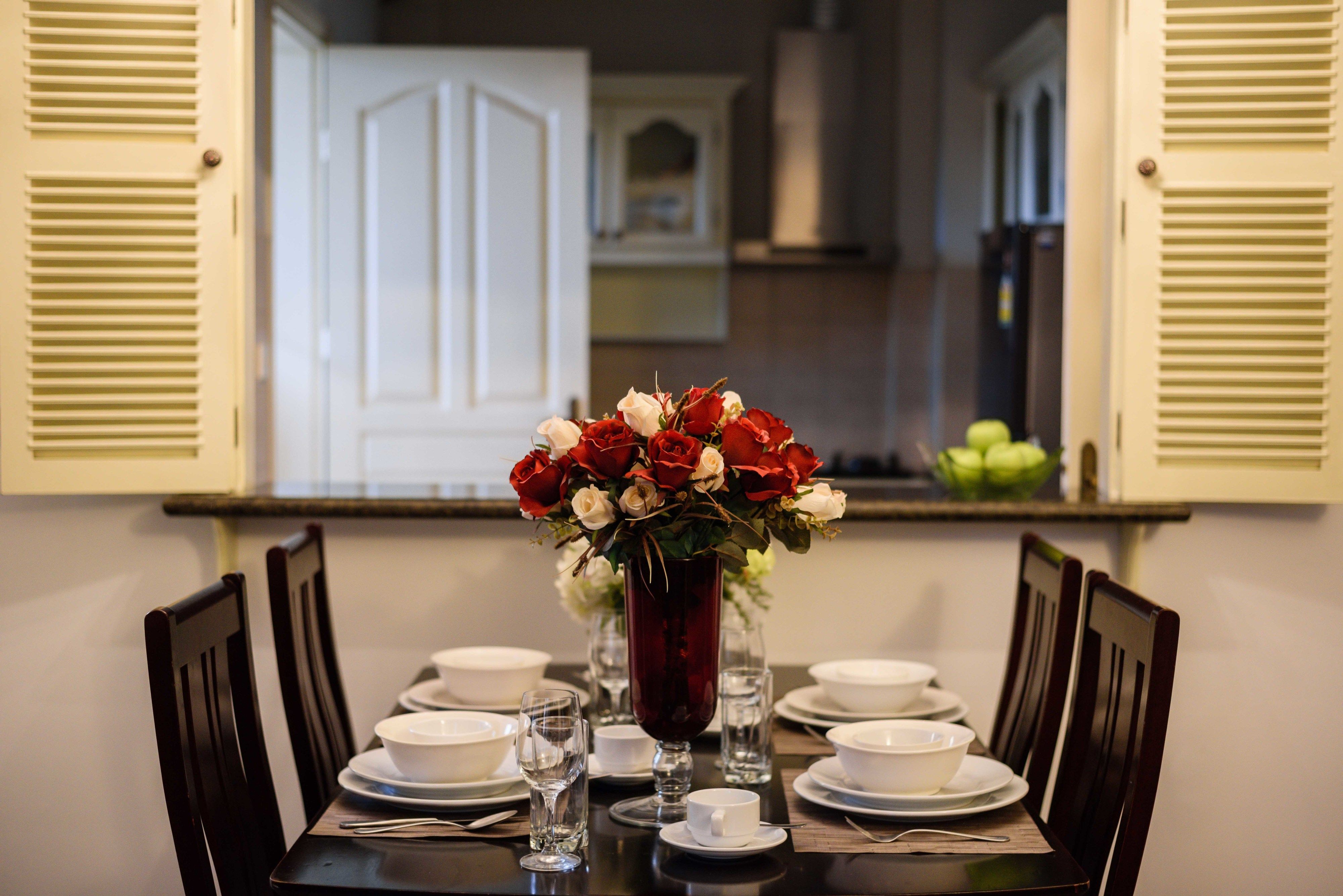 Een eettafel is gedekt met witte borden, kommen en glazen, en in het midden staat een vaas met rode en witte bloemen.
