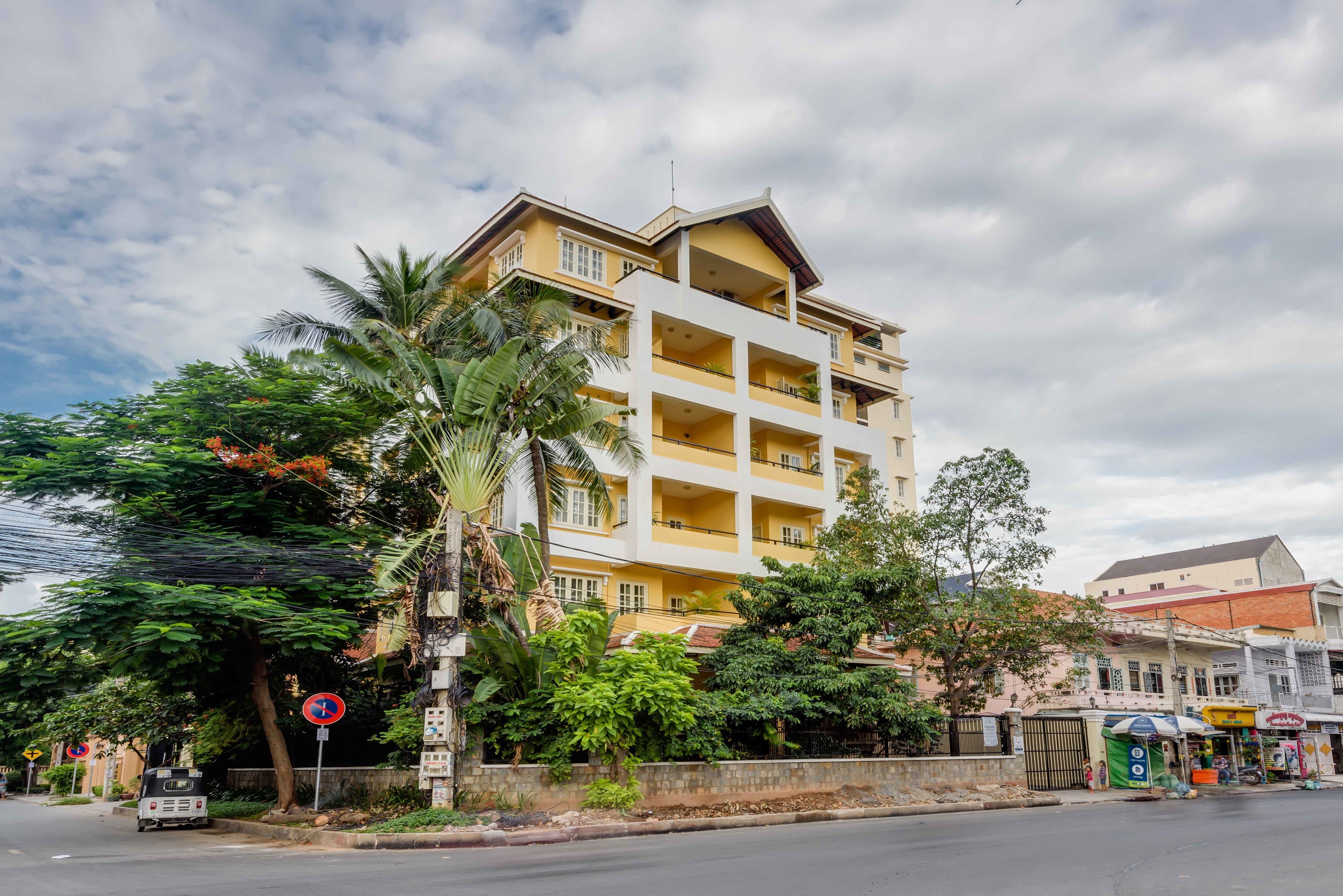 A multi-story building with balconies, surrounded by greenery and a street.