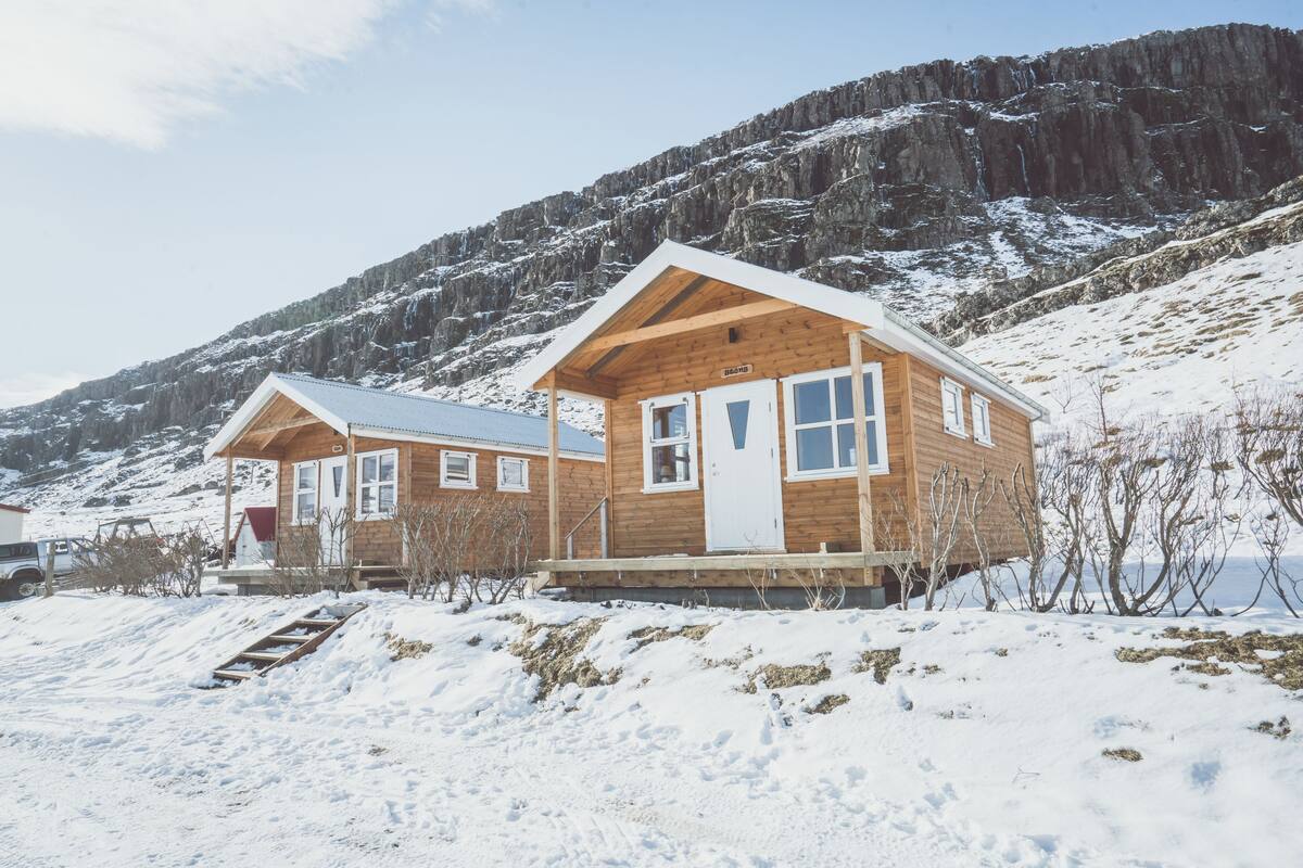 A wooden cabin with a white door and windows, set against a snowy mountain backdrop.