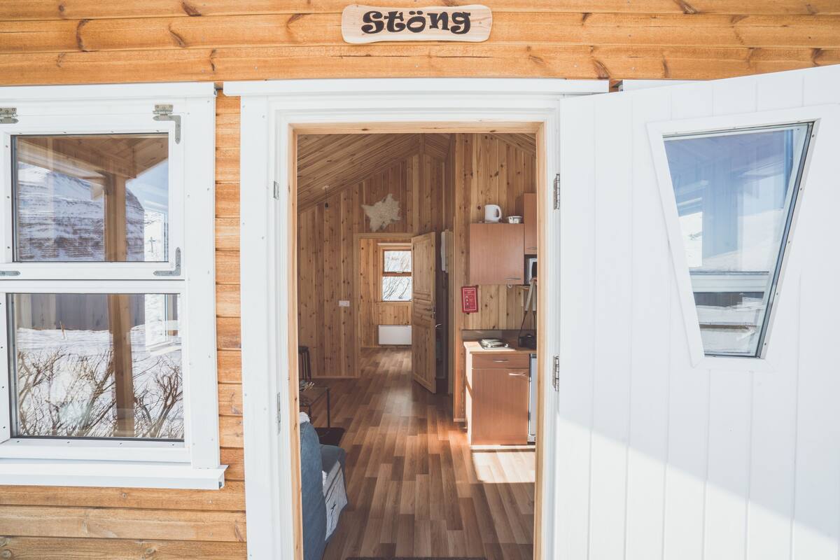 A wooden cabin interior with a visible kitchen area, a dining table, and a sign reading "Stöng".