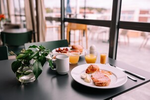 Una mesa de desayuno con un croissant, jamón, queso, jugo, un florero con hojas verdes y una taza.