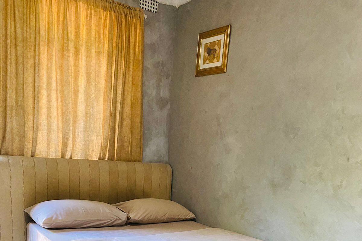 Ein Einzelbett mit beiger Bettdecke und Kissen, ein Kopfteil, ein gerahmtes Bild an der Wand und ein Fenster mit Vorhängen.