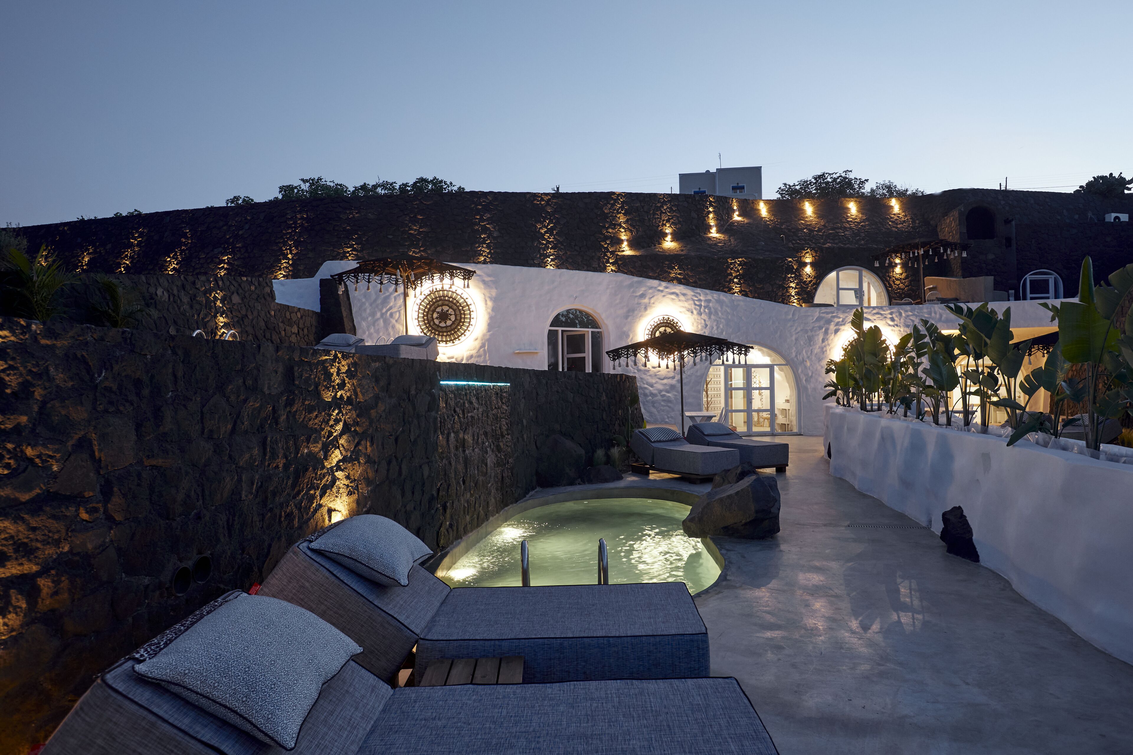 A rooftop pool area with lounge chairs and a hot tub, surrounded by stone walls and plants.