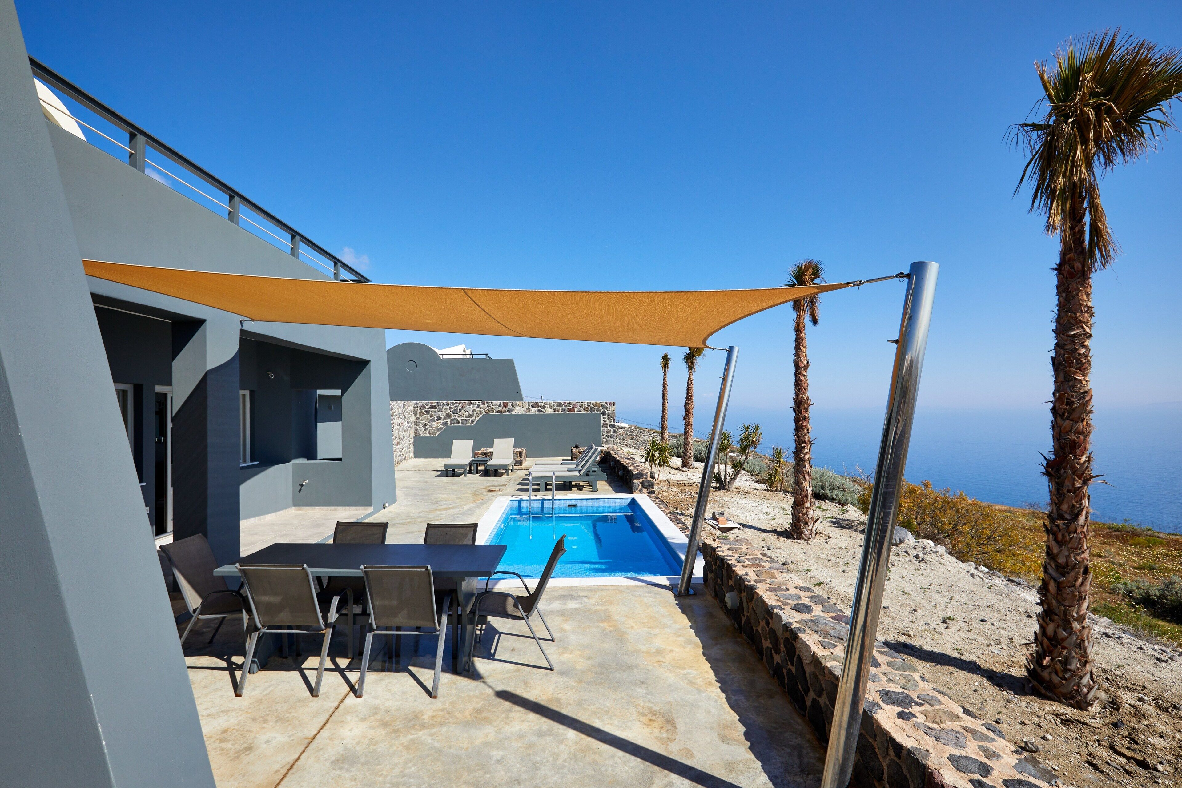 A modern outdoor pool area with a stone wall, a table with chairs, and a view of the sea.