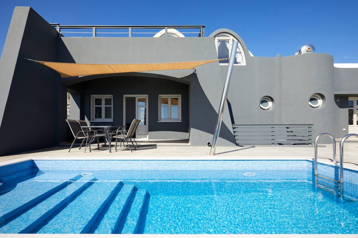 A swimming pool with a sunshade, a seating area with chairs, and a modern building with large windows.