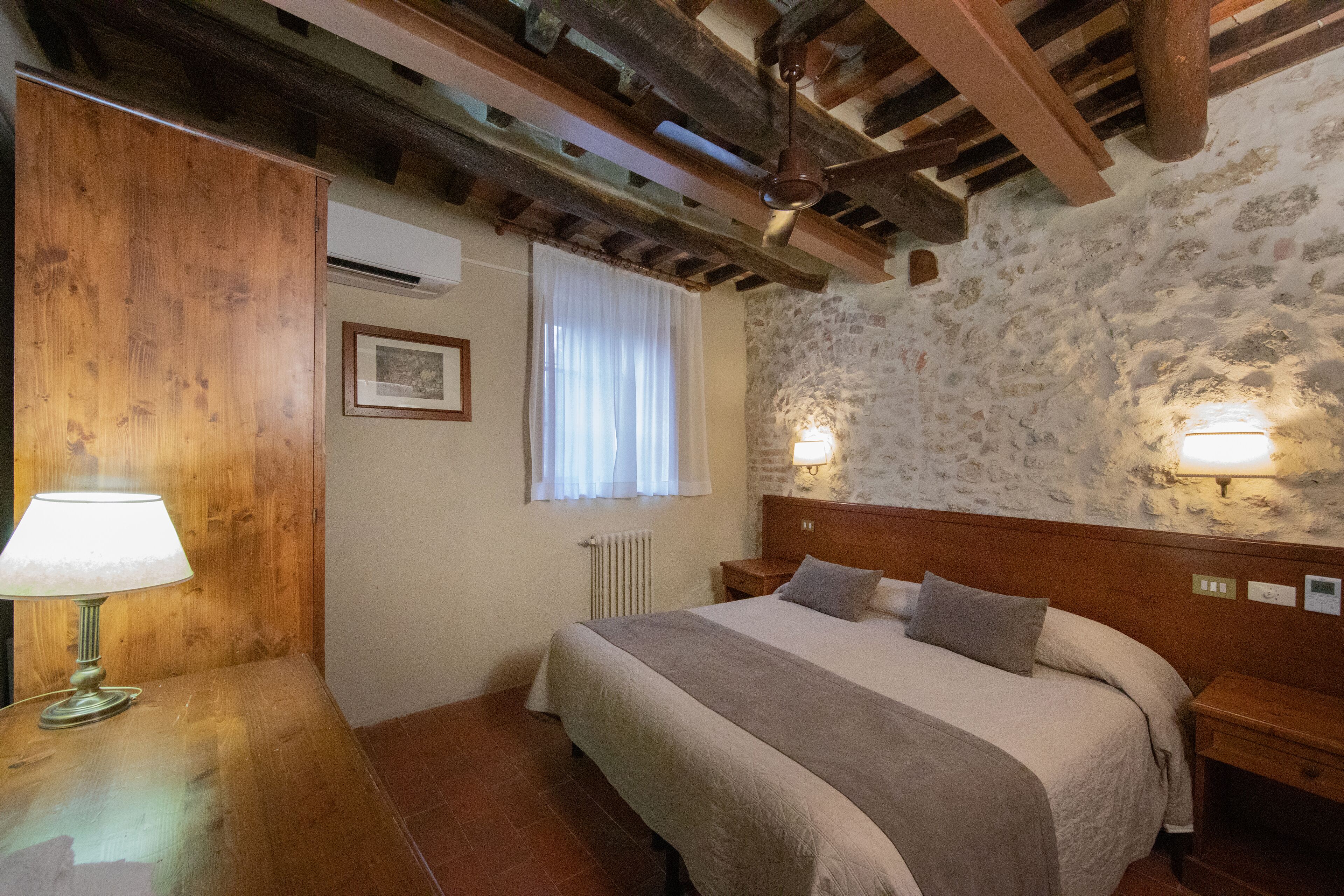 A bedroom with a wooden ceiling, stone walls, a bed with a headboard, bedside lamps, and a radiator.