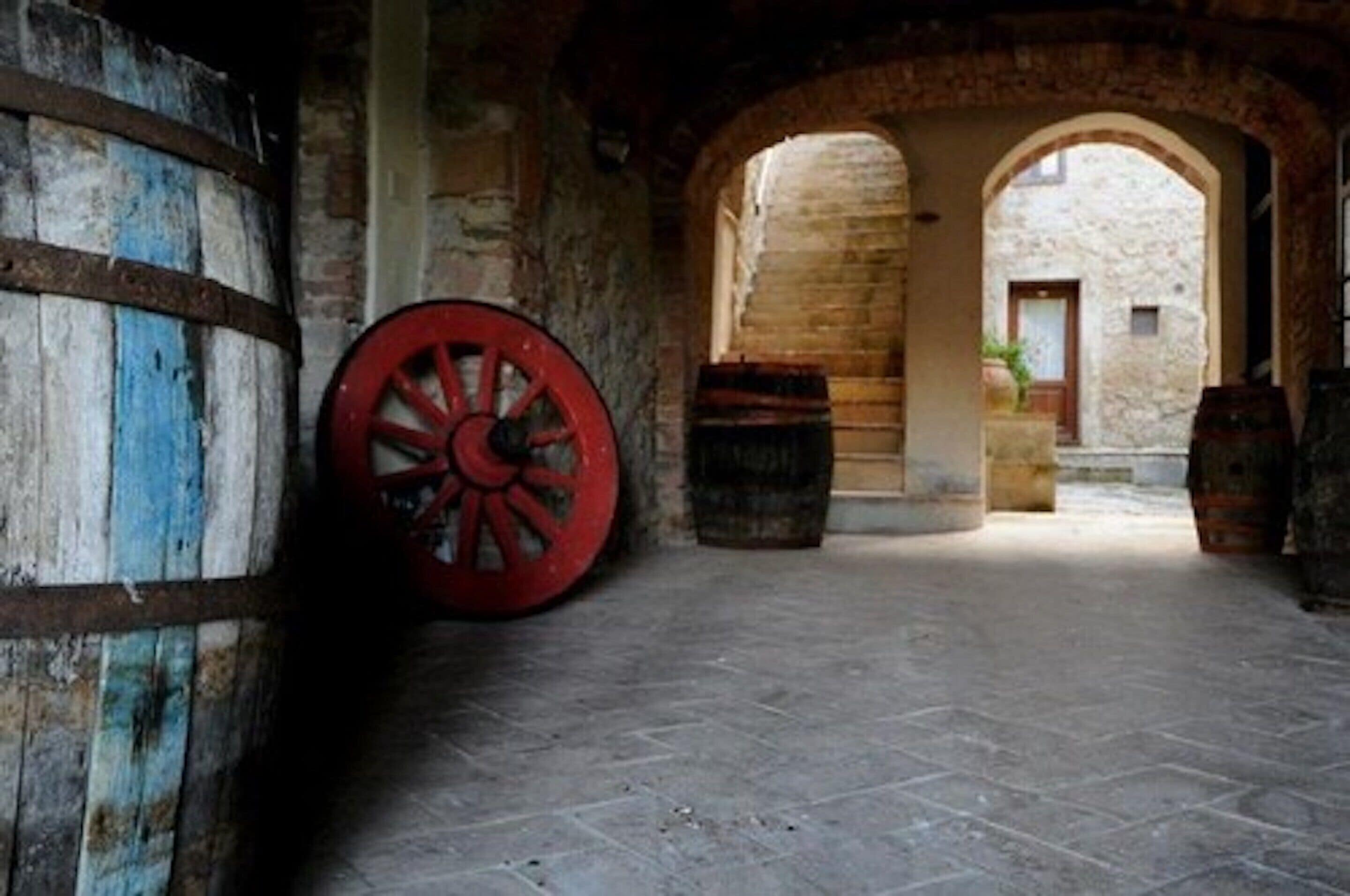 Un couloir en pierre, avec un grand tonneau en bois et une roue rouge, qui mène à une porte en arc de cercle.