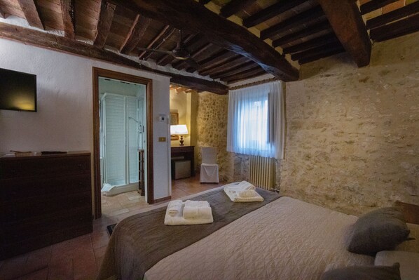 A hotel room with a wooden ceiling, a bed with a quilt and pillows, a bedside table, a lamp, and a view of a stone wall. - Tenuta Decimo (San Gimignano)