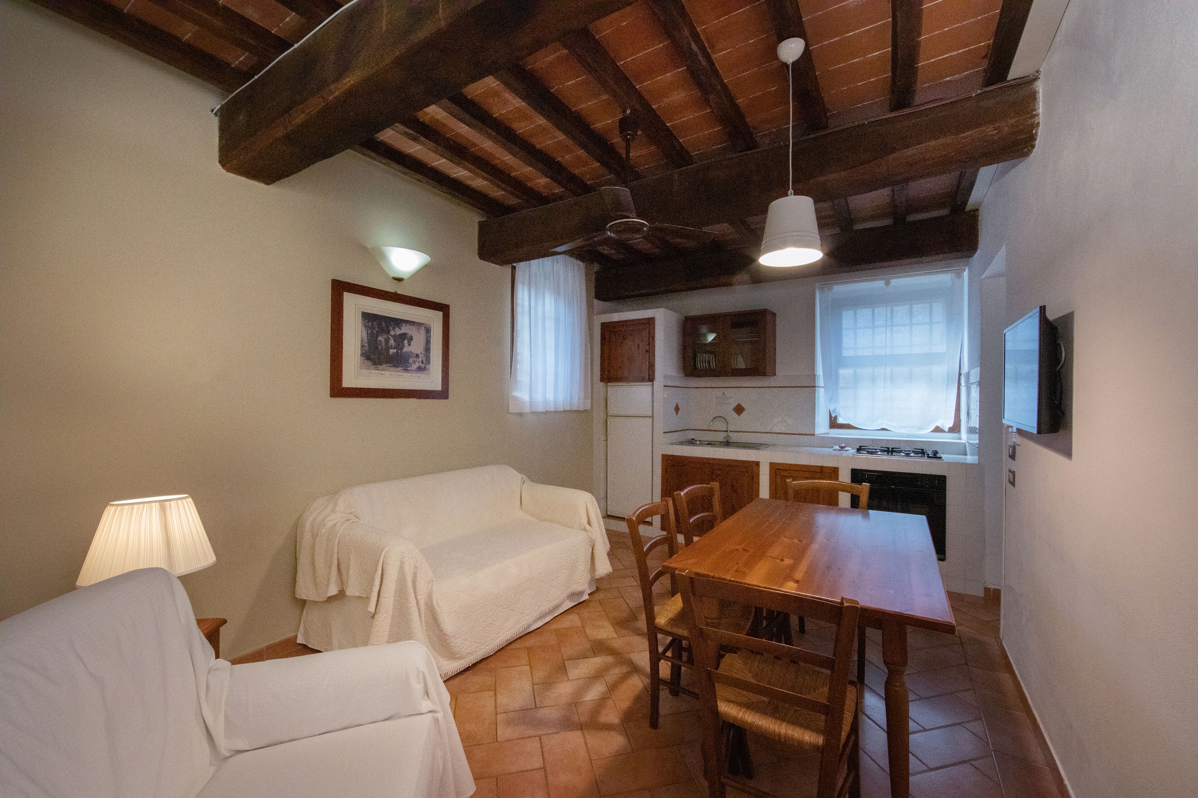 Una camera accogliente con un tavolo da pranzo in legno, due sedie, un divano, una lampada, una ventola a soffitto e un angolo cottura con una finestra.