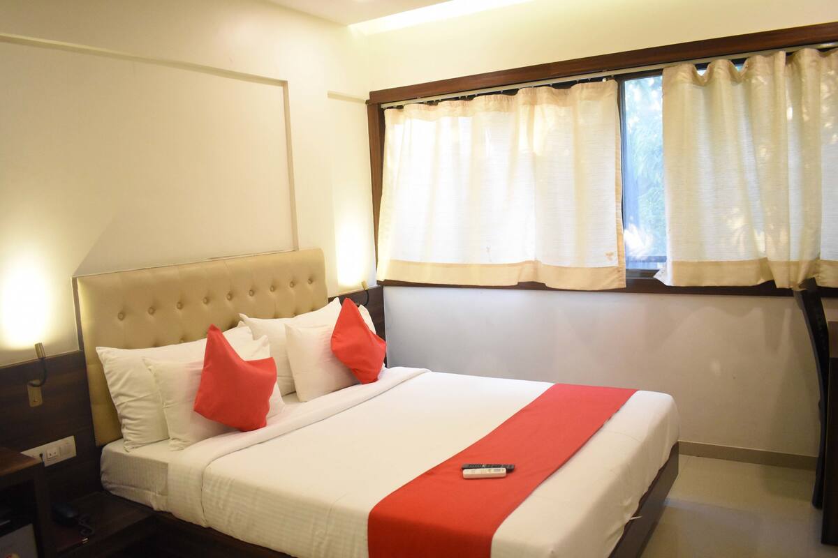 Une chambre d’hôtel avec un grand lit, du linge de lit blanc, des touches de couleur rouge, une télécommande et une fenêtre avec des rideaux.