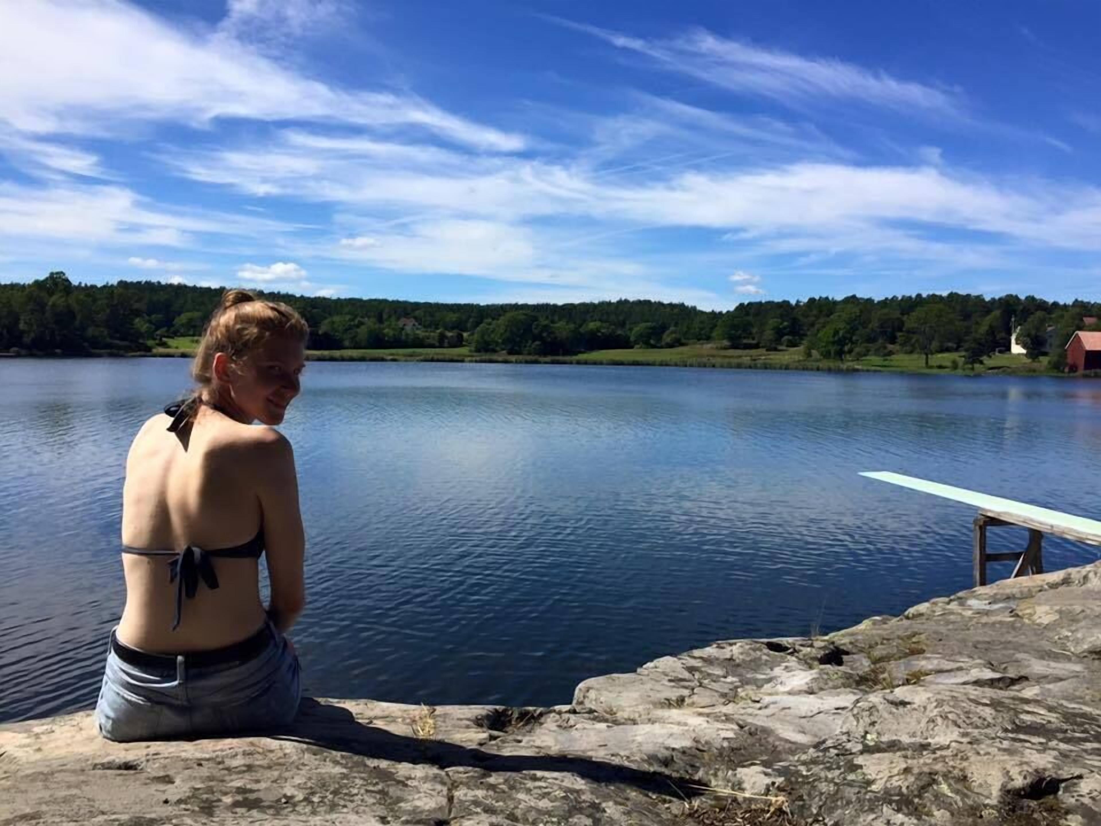 Una persona sentada sobre un saliente rocoso junto a un lago tranquilo con un muelle de madera que se adentra en el agua.