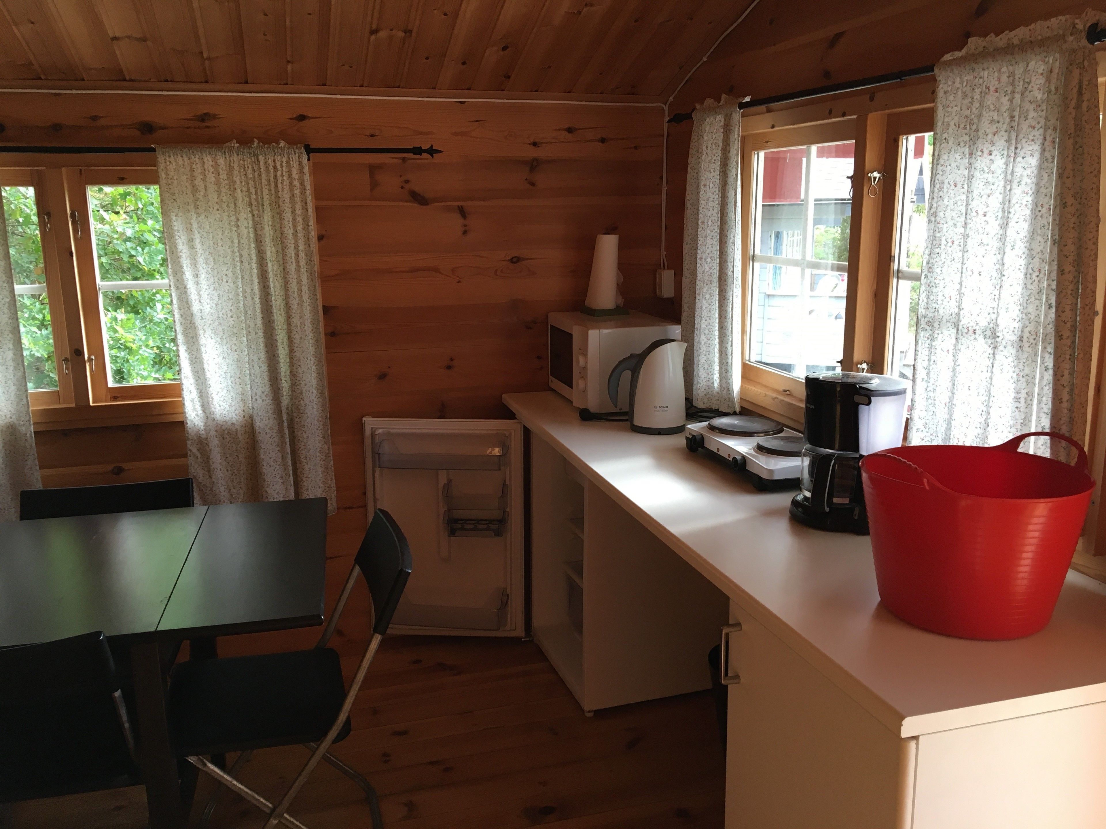 A compact kitchen with wooden walls, a small refrigerator, a microwave, a kettle, and a red bucket on the counter.