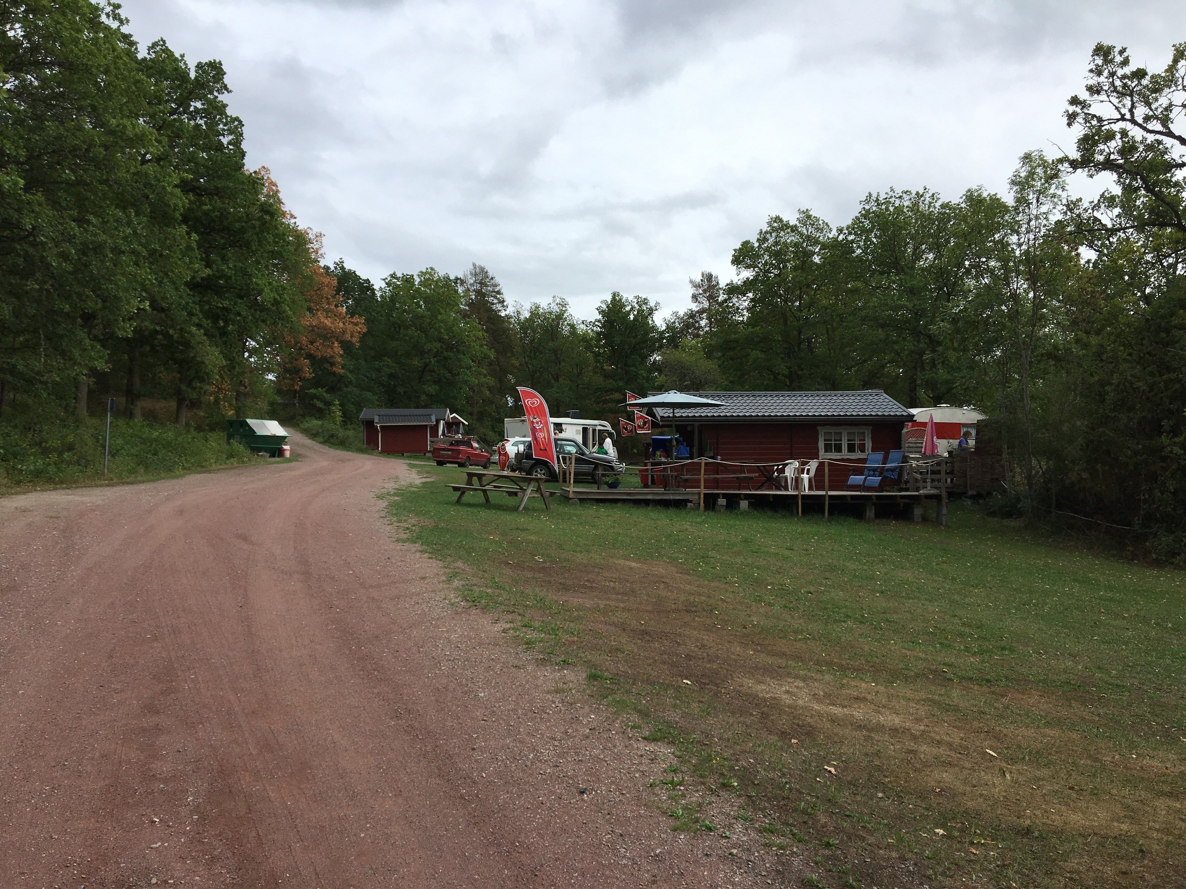 A campground with a dirt road leading to a red building, picnic tables, and a covered area with seating.