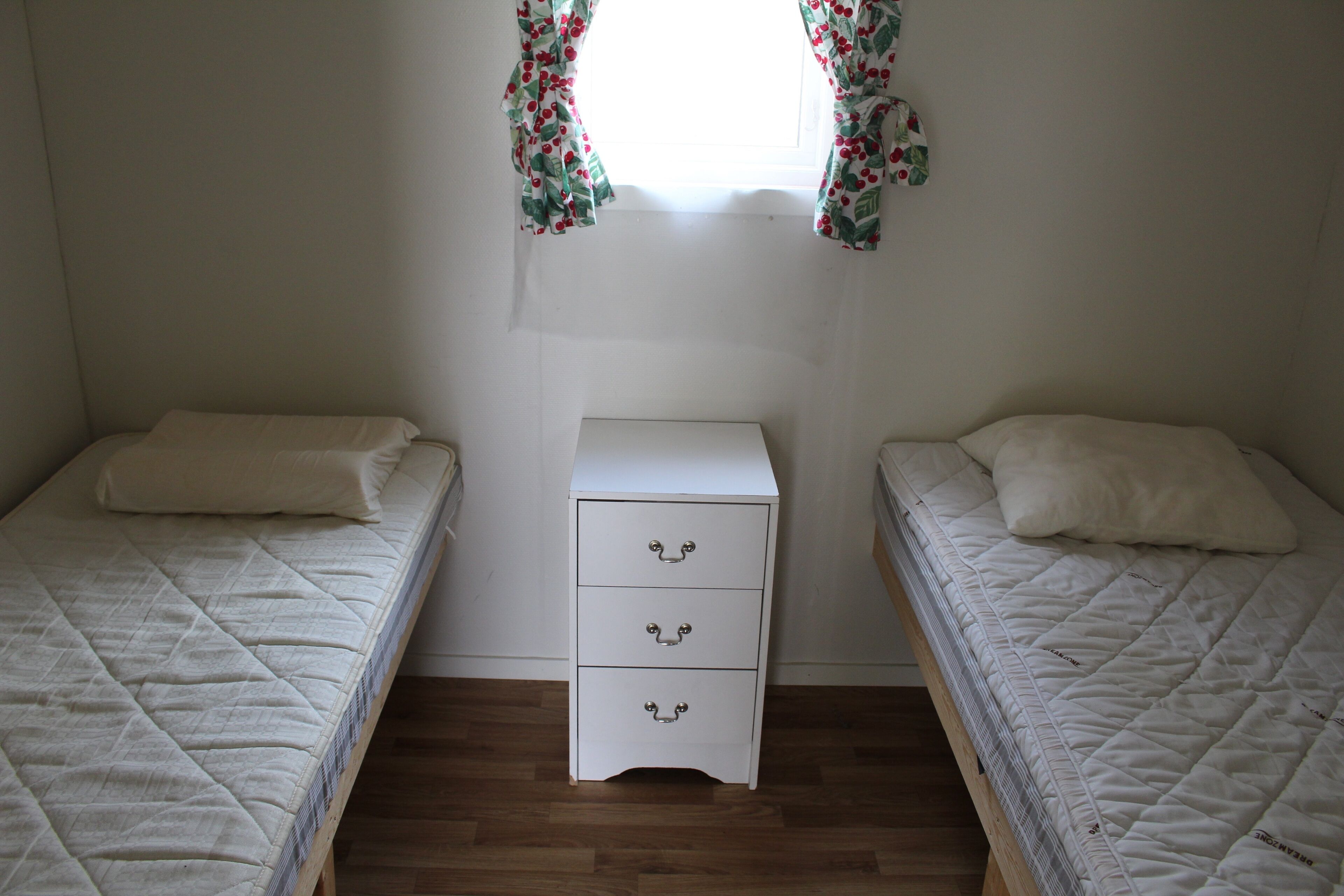 A small, simple room with two single beds, a white nightstand with drawers, and a window with floral curtains.
