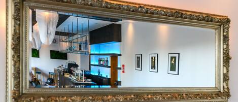 A mirror with a gold frame reflecting a bar area with shelves of bottles, a staircase, and a ceiling light fixture.