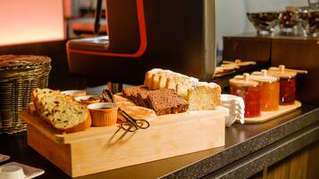 A variety of breads and pastries on a wooden board, with a coffee machine and jars of jam in the background.