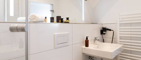 Un baño moderno con inodoro blanco, lavamanos y ducha. Hay un perchero y un espejo sobre el lavamanos.