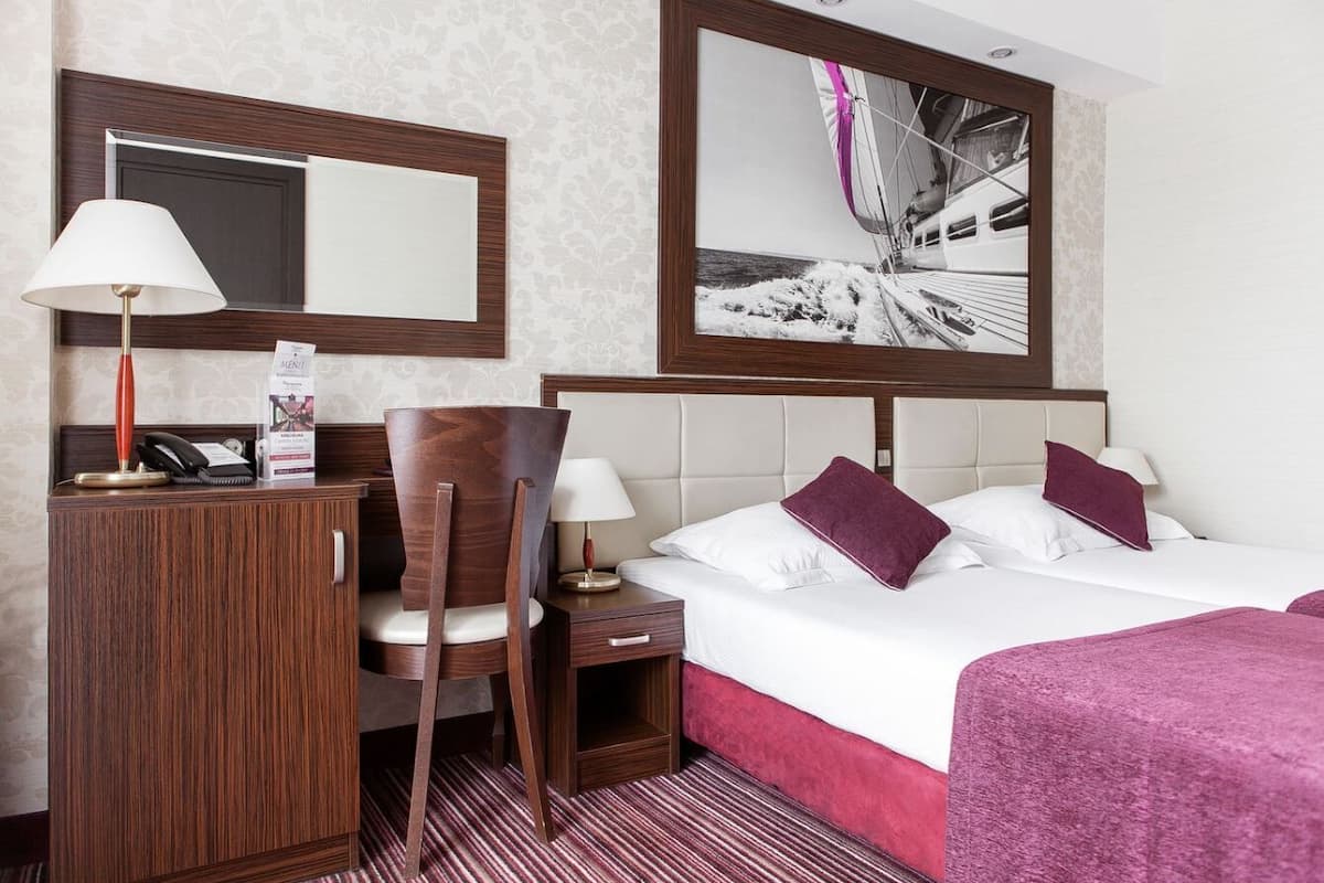 A hotel room with a bed, a desk, a chair, a nightstand, a lamp, a mirror, and a framed picture on the wall.
